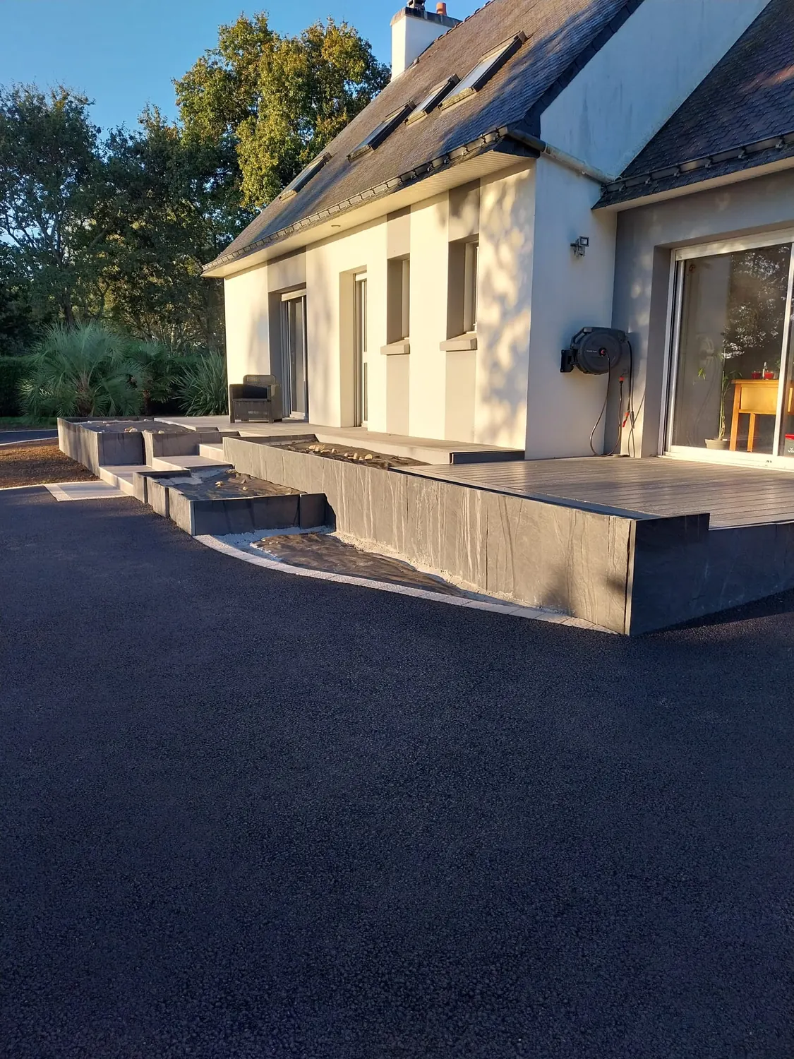 Cour en enrobé au pied d'une terrasse béton, lumière du soir